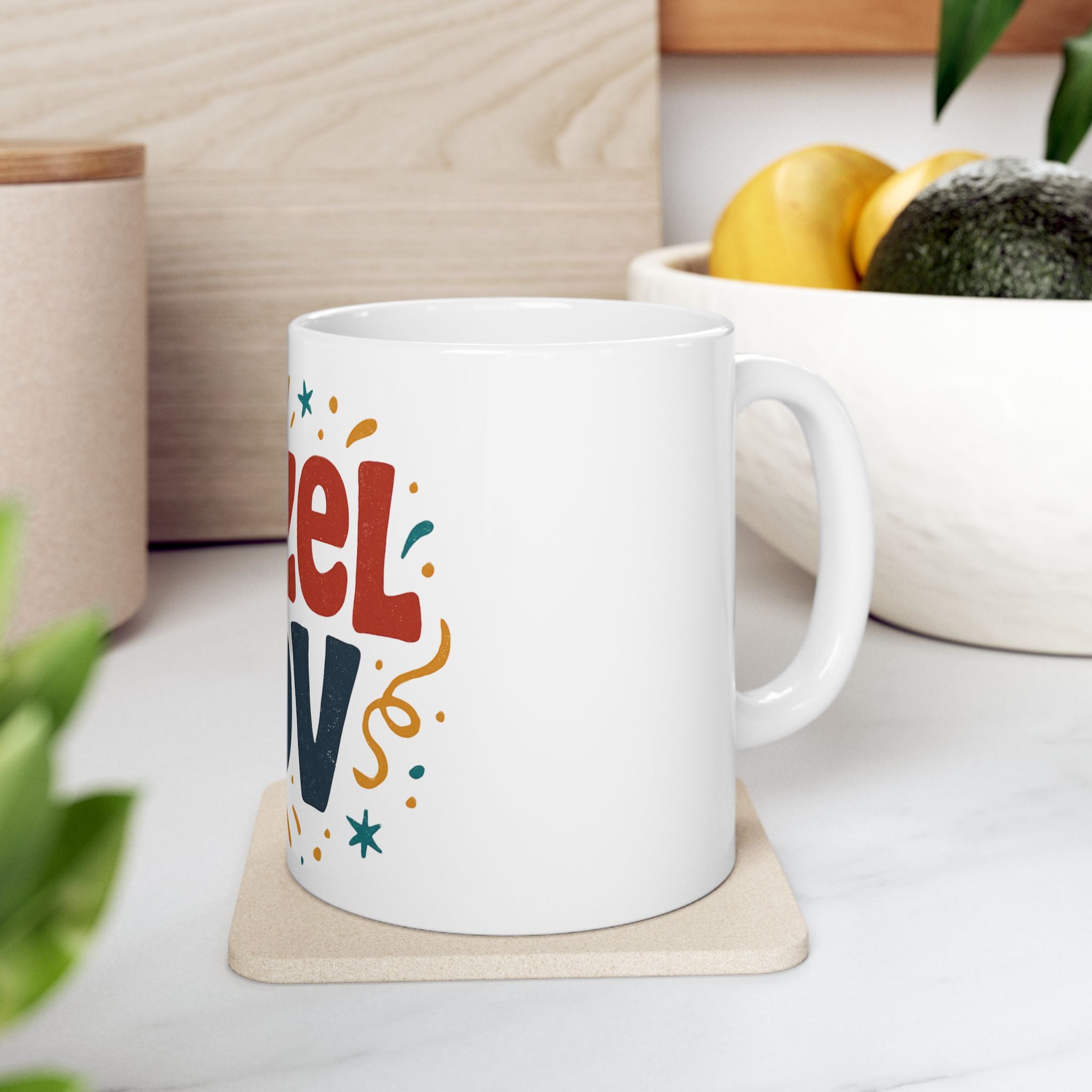 White mug with colorful text on a coaster, placed on a surface with a plant and container in the background.