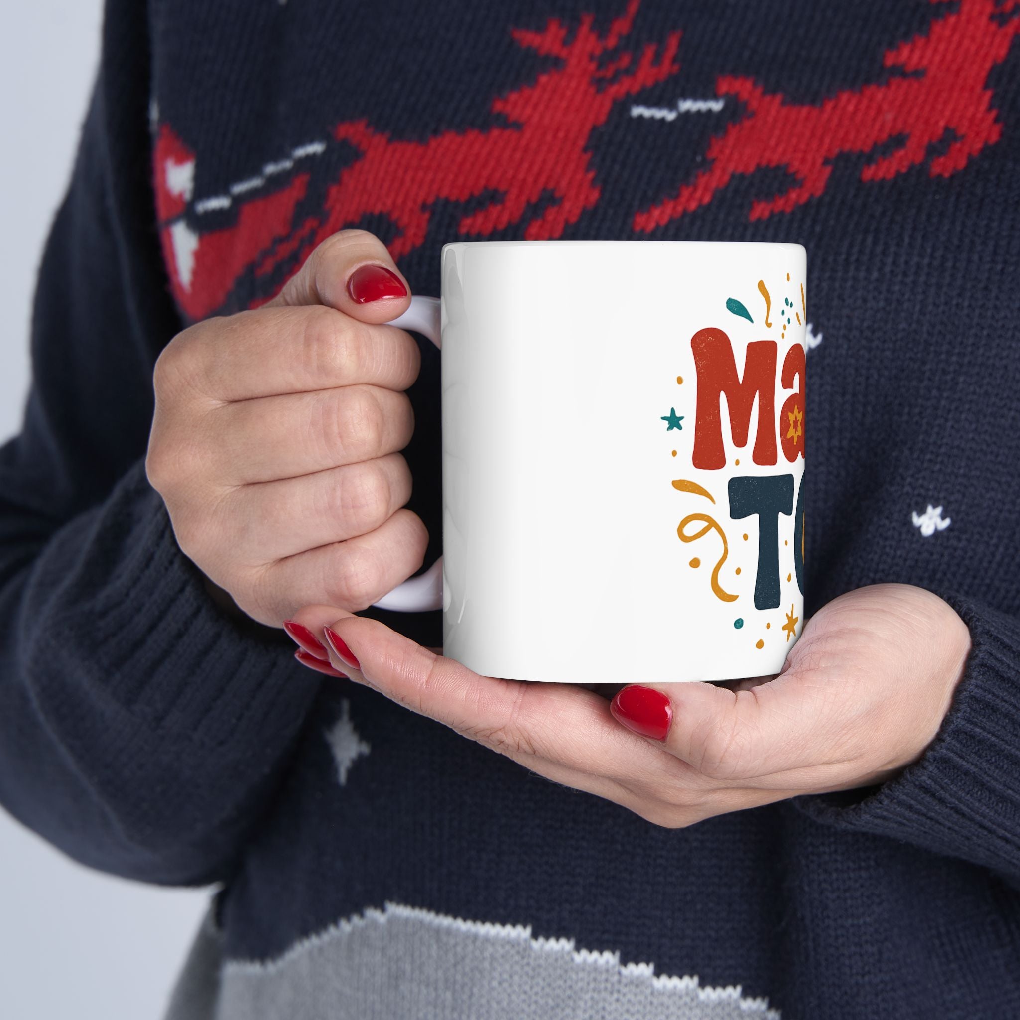 Person holding a white mug with colorful text, wearing a dark sweater with red deer designs.
