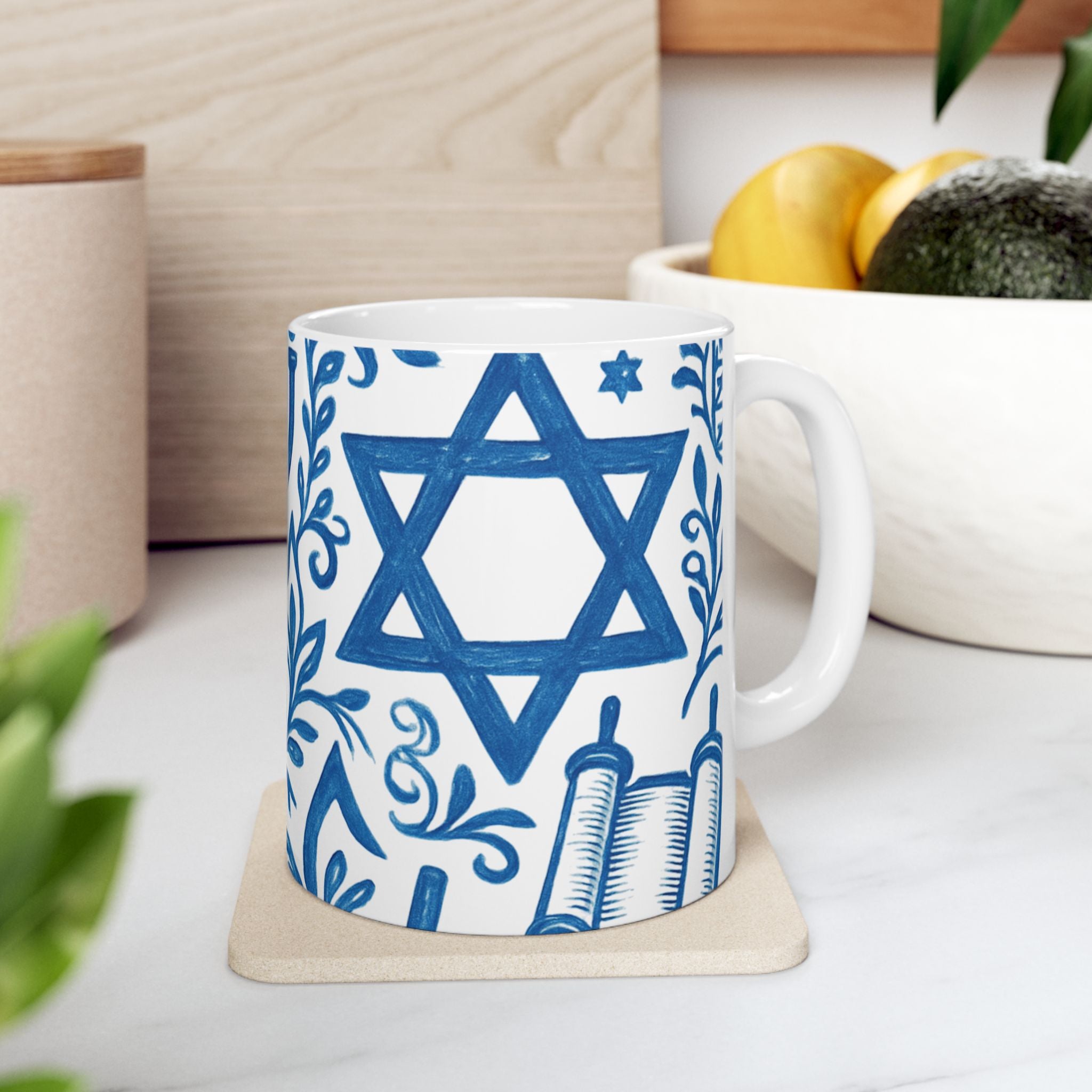 White mug with blue Star of David and religious symbols on a white surface with plants and a plant pot in the background.